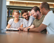 Vier Personen sitzen an einem Tisch in der Bibliothek der Hochschule Coburg, umgeben von Bücherregalen. Sie lächeln und unterhalten sich, vor ihnen liegt ein aufgeklappter Laptop. Die Umgebung wirkt hell und lädt zum Lernen oder Zusammenarbeiten ein.