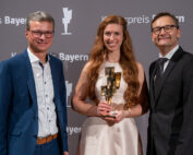 Drei Menschen stehen lächelnd bei einer Preisverleihung zusammen. Die Frau in der Mitte, Vertreterin der Hochschule Coburg, hält eine Trophäe in der Hand. Sie sind in formelle Kleidung gekleidet und stehen vor einem Hintergrund mit der Aufschrift „Kulturpreis Bayern“.