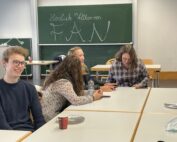 Eine Gruppe von vier Personen sitzt an Tischen in einem Hörsaal, vor ihnen stehen Tassen und Teller. Im Hintergrund steht auf einer Tafel „Herzlich Willkommen FAN, Hochschule Coburg“. Alle scheinen in ein Gespräch vertieft zu sein.