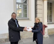 Zwei Menschen lächeln und halten gemeinsam eine weiße Auszeichnungsplakette vor einem Gebäude mit Säulen und Bogenfenstern der Hochschule Coburg. Die Person links trägt einen schwarzen Mantel, die Person rechts trägt eine Brille, eine dunkle Jacke und einen roten Rucksack.