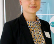 Eine Person mit hellbraunem Haar, einem schwarzen Blazer und einer gemusterten Bluse steht selbstbewusst vor einer Tafel mit Text an der Hochschule Coburg.