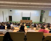 In einem überfüllten Hörsaal der Hochschule Coburg herrscht reges Treiben. Studenten sitzen vor einem Dozenten. Der Dozent steht an einem Rednerpult vor einer großen Tafel. Auf zwei Bildschirmen läuft eine Präsentation. Der Raum ist hell erleuchtet, die Holztische stehen in Reihen, die Wände sind weiß.