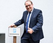 Ein Mann in Anzug und Krawatte steht neben einem Holzpfosten mit einem Schild, auf dem „Hochschule Coburg“ und „Hochschulleitung“ stehen. Er lächelt und zeigt mit der linken Hand den Daumen nach oben. Der Hintergrund ist eine schlichte weiße Wand.