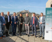 Eine Gruppe von sieben Personen posiert auf einem Balkon mit malerischer Aussicht auf eine Burg auf einem Hügel. Sie stehen neben einem Werbebanner für das „Promotionszentrum Nachhaltige und Intelligente Systeme (NISys)“ der Hochschule Coburg.