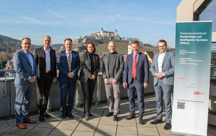 Eine Gruppe von sieben Personen posiert auf einem Balkon mit malerischer Aussicht auf eine Burg auf einem Hügel. Sie stehen neben einem Werbebanner für das „Promotionszentrum Nachhaltige und Intelligente Systeme (NISys)“ der Hochschule Coburg.