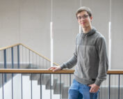 Eine Person mit Brille und grauem Pullover lehnt an einem Geländer in einem modernen Gebäude der Hochschule Coburg, im Hintergrund windet sich eine Treppe elegant.