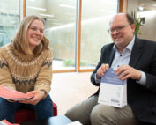 Zwei Menschen sitzen in einer modernen Bürolounge der Hochschule Coburg, lächeln und halten Bücher und Papiere in den Händen. Einer trägt einen Strickpullover, der andere einen Anzug. Sie sitzen auf roten Stühlen, im Hintergrund sind große Fenster zu sehen.