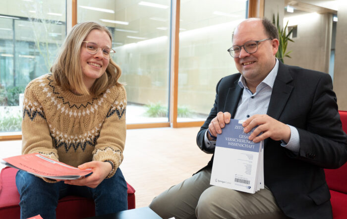 Zwei Menschen sitzen in einer modernen Bürolounge der Hochschule Coburg, lächeln und halten Bücher und Papiere in den Händen. Einer trägt einen Strickpullover, der andere einen Anzug. Sie sitzen auf roten Stühlen, im Hintergrund sind große Fenster zu sehen.