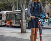 Eine Frau, vermutlich eine Studentin der Hochschule Coburg, fährt mit einem roten Elektroroller eine Stadtstraße entlang. Sie trägt einen Helm und ein blaues Outfit mit einer gemusterten Jacke. Im Hintergrund, während sie an dem von Bäumen gesäumten Bürgersteig vorbeifährt, rollt ein Bus vorbei.