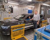 Ein Mann steht neben einem Auto mit der Aufschrift „Synfuel“ auf dem Testgelände der Hochschule Coburg. Eine weitere Person sitzt im Auto. Der Raum ist voller Maschinen und Bedienfelder, die die wissenschaftliche und ingenieurtechnische Kompetenz der Hochschule unterstreichen.