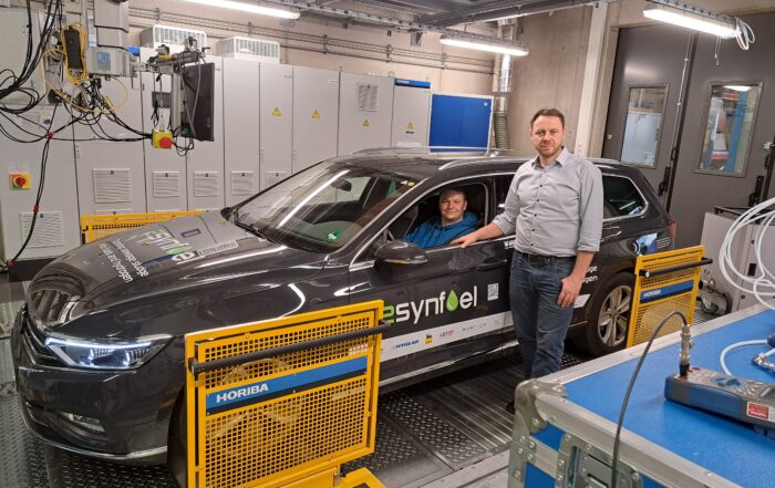 Ein Mann steht neben einem Auto mit der Aufschrift „Synfuel“ auf dem Testgelände der Hochschule Coburg. Eine weitere Person sitzt im Auto. Der Raum ist voller Maschinen und Bedienfelder, die die wissenschaftliche und ingenieurtechnische Kompetenz der Hochschule unterstreichen.