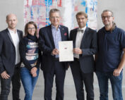 Fünf Personen stehen zusammen im Innenbereich und lächeln strahlend. Die zentrale Figur hält stolz ein Zertifikat der Hochschule Coburg in den Händen. Sie sind in Business- und Freizeitkleidung gekleidet, ein modernes, abstraktes Gemälde unterstreicht die Atmosphäre im Hintergrund.