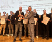 Eine Gruppe von Mitarbeitern der Hochschule Coburg steht mit Zertifikaten auf der Bühne. Zwei Männer in der ersten Reihe schütteln einander die Hände und tauschen einen Pokal aus. Im Hintergrund ist eine Präsentationsfolie mit Text und einer Grafik zu sehen.