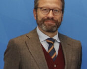 Vor einem blauen Hintergrund steht ein Mann mit Brille, Vollbart und Schnurrbart. Er trägt ein braunes Jackett über einer roten Pullunderweste und einer gestreiften Krawatte. Sein selbstbewusster Blick spiegelt die akademische Exzellenz wider, die mit der Hochschule Coburg verbunden wird.