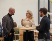 Drei Personen diskutieren an einem Holztisch mit weißen Kaffeetassen in einem Büro der Hochschule Coburg. Die Frau in der Mitte gestikuliert mit den Händen, während sie spricht, und die beiden Männer hören aufmerksam zu.