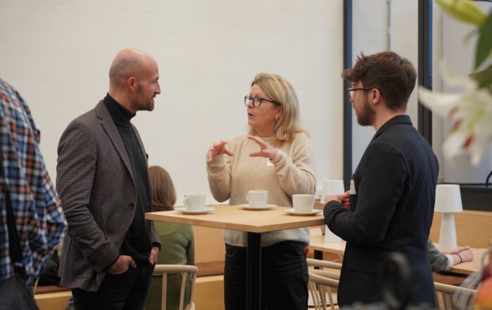 Drei Personen diskutieren an einem Holztisch mit weißen Kaffeetassen in einem Büro der Hochschule Coburg. Die Frau in der Mitte gestikuliert mit den Händen, während sie spricht, und die beiden Männer hören aufmerksam zu.