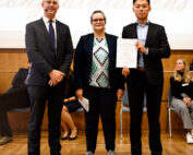 Drei Personen stehen auf einer Bühne. Zwei von ihnen halten Zertifikate in den Händen. Links steht ein Mann im Anzug, daneben stehen eine Frau und ein weiterer Mann, beide in formeller Kleidung. Hinter ihnen wird „Herzlichen Glückwunsch“ projiziert und das Logo der Hochschule Coburg.