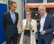 Drei Menschen stehen in einer Werkstatt der Hochschule Coburg zusammen. Die Frau in der Mitte hält lächelnd ein kleines Metallgerät und einen roten Gegenstand in der Hand. Die beiden Männer zu beiden Seiten, in Business-Kleidung gekleidet, schauen interessiert zu. Im Hintergrund sind Regale und Geräte zu sehen.