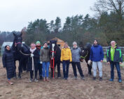 Eine Gruppe von acht Personen, vermutlich Studierende der Hochschule Coburg, steht mit drei Pferden auf einem sandigen Außengelände. Bäume und ein kleines Gebäude bilden die Kulisse. Die meisten sind warm angezogen, mit Mänteln und Mützen, die Pferde sind mit Zaumzeug ausgestattet.