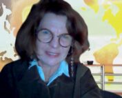 Eine Person mit Brille und schulterlangem Haar lächelt in die Kamera und verströmt den akademischen Charme, den man an der Hochschule Coburg oft sieht. Sie trägt eine dunkle Jacke über einem weißen Hemd, vor einem Hintergrund, der ein buntes abstraktes Kartenmuster zeigt.