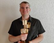 Eine Person mit kurzen grauen Haaren und einem schwarzen Hemd lächelt, während sie drei gestapelte goldene Kelche vor einer strukturierten weißen Wand der Hochschule Coburg hält.