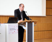 Ein Mann im Anzug steht an einem Podium in einem Konferenzsaal und spricht in ein Mikrofon. Auf dem Podium stehen ein Laptop und ein Glas Wasser. Auf einem Plakat steht „Erster Coburger Mobilitätskongress“, stolz präsentiert von der Hochschule Coburg.