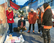 Eine Gruppe von sechs Menschen in warmer Kleidung und mit Masken versammelt sich auf einer Kopfsteinpflasterstraße vor der Hochschule Coburg und beobachtet eine Person, die in der Hocke sitzt und auf Plakate mit den Aufschriften „STOP WAR“ und „PAY“ zeigt. Es ist eine Abendszene in einer europäischen Stadt, mit Geschäften und beleuchteten Gebäuden im Hintergrund.