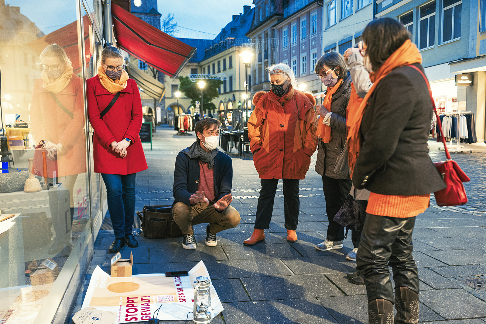 Eine Gruppe von sechs Menschen in warmer Kleidung und mit Masken versammelt sich auf einer Kopfsteinpflasterstraße vor der Hochschule Coburg und beobachtet eine Person, die in der Hocke sitzt und auf Plakate mit den Aufschriften „STOP WAR“ und „PAY“ zeigt. Es ist eine Abendszene in einer europäischen Stadt, mit Geschäften und beleuchteten Gebäuden im Hintergrund.