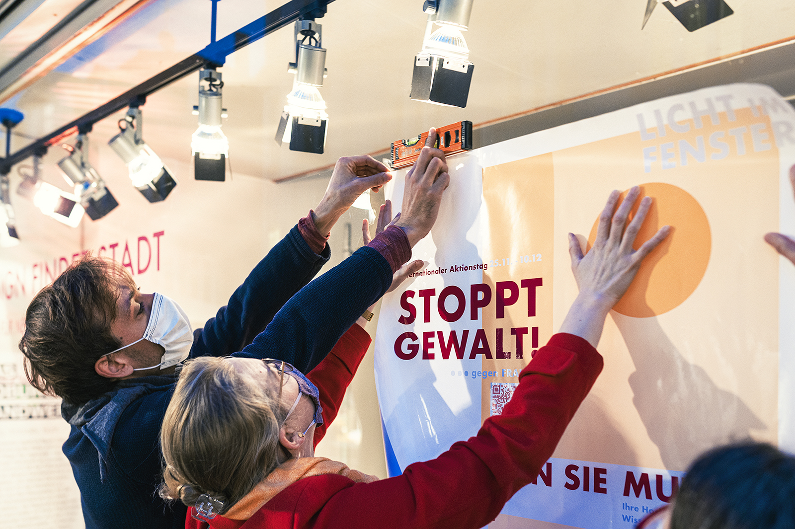 Zwei Personen, eine davon mit Mundschutz, richten vorsichtig ein großes Plakat an einer Wand der Hochschule Coburg aus. Auf dem Plakat steht „Stoppt Gewalt!“. Mit einer Wasserwaage stellen sie sicher, dass es unter den hellen Deckenleuchten gerade hängt.