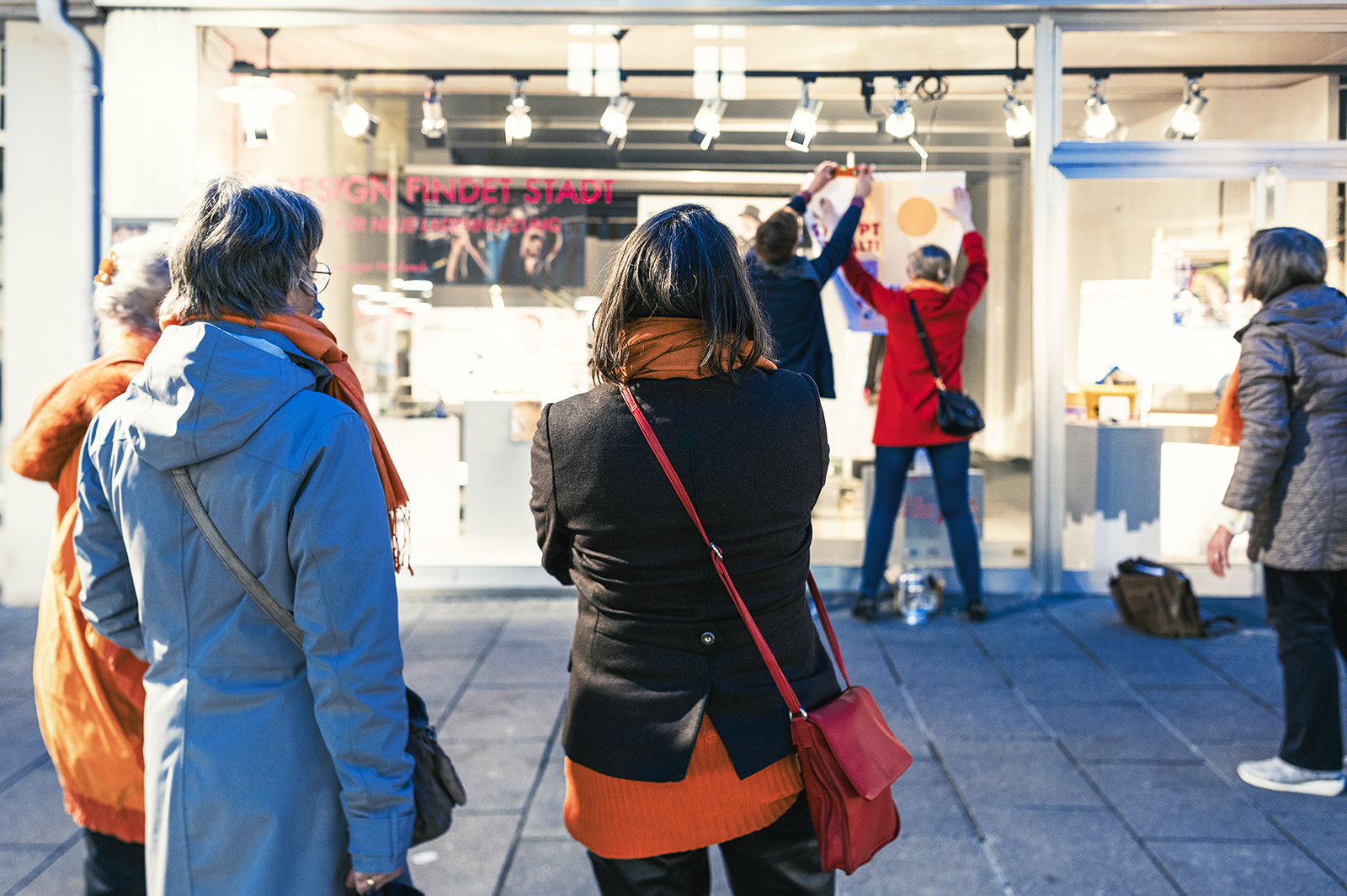 Eine Gruppe von Menschen steht nachts vor einem Schaufenster der Hochschule Coburg und beobachtet, wie im Inneren eine Ausstellung aufgebaut wird. Zwei Personen arbeiten an der Anordnung von Kunstwerken, während andere zuschauen. Die Straße ist hell erleuchtet und die Szenerie ist von lebhafter Atmosphäre geprägt.