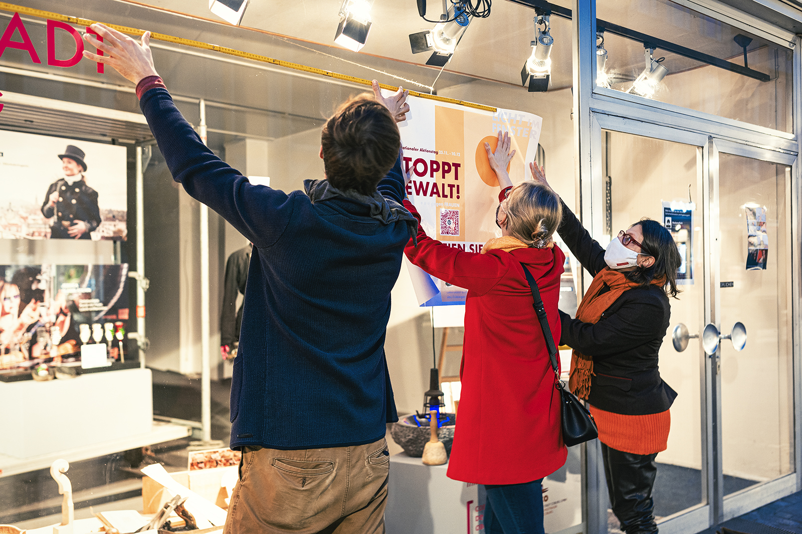 Drei Personen arbeiten zusammen, um ein Plakat an das Glasfenster der Hochschule Coburg zu hängen. Einer misst, während ein anderer das Plakat hält, alle in Jacken und Schals eingepackt. Durch das Glas sind verschiedene Objekte und Lichter im Inneren zu sehen, die auf das pulsierende akademische Leben im Inneren hinweisen.