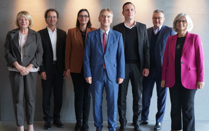 Eine Gruppe von sieben Personen steht in einem Innenbereich der Hochschule Coburg vor einer grauen Wand. Fünf von ihnen sind in professioneller Kleidung gekleidet, fünf tragen Sakko über Hemden, während zwei Frauen leuchtende Blazer tragen – einer rosa, einer orange – und selbstbewusst in die Kamera blicken.