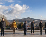 Eine Gruppe von acht Personen steht auf einer breiten Steinplattform vor einer malerischen Kulisse aus Hügeln, Bäumen und dem Schloss in der Ferne in der Nähe der Hochschule Coburg. Der Himmel ist teilweise bewölkt und das Herbstlaub verleiht der Landschaft leuchtende Farben.