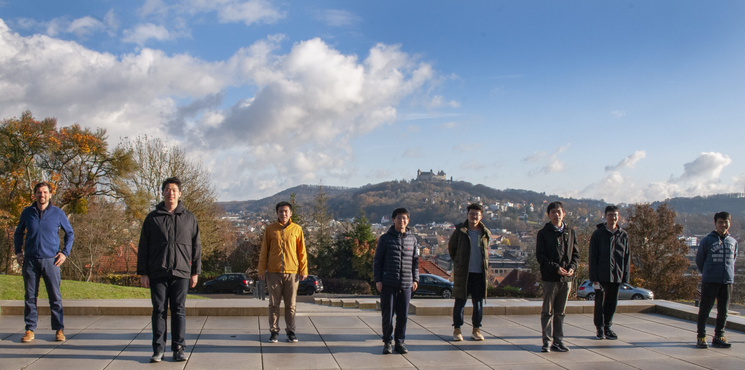 Eine Gruppe von acht Personen steht auf einer breiten Steinplattform vor einer malerischen Kulisse aus Hügeln, Bäumen und dem Schloss in der Ferne in der Nähe der Hochschule Coburg. Der Himmel ist teilweise bewölkt und das Herbstlaub verleiht der Landschaft leuchtende Farben.