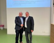 Zwei Männer stehen lächelnd vor einem Präsentationsbildschirm in der Hochschule Coburg und schütteln sich die Hände. Beide tragen Anzüge und passen so gut zum grünen Boden und den weißen Wänden des Raums. Eine teilweise sichtbare Folie trägt zur professionellen Atmosphäre bei.