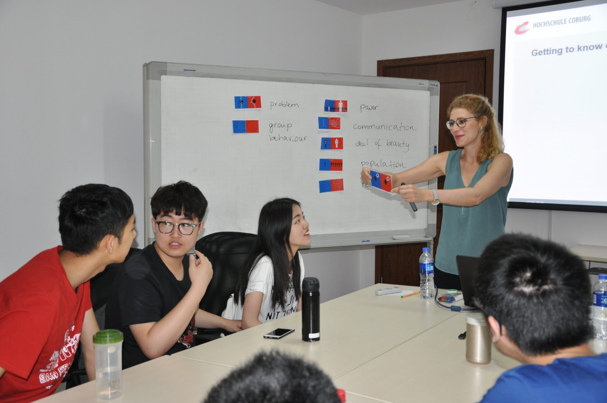 Eine Frau zeigt auf eine Flaggentafel und erklärt einer Gruppe engagierter Studierender, die an einem Tisch der Hochschule Coburg sitzen, etwas. Die Studierenden hören aufmerksam zu, und im Raum herrscht eine kollaborative Atmosphäre.
