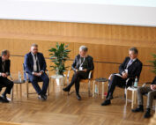 Fünf Personen sitzen im Halbkreis auf der Bühne mit Holzboden der Hochschule Coburg und diskutieren an einem Podium. Sie halten Mikrofone in den Händen und haben Wasserflaschen bereitgelegt. Topfpflanzen schmücken den Hintergrund, über ihnen ist eine große Projektionsfläche zu sehen.