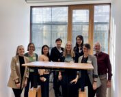 Eine Gruppe von acht Personen der Hochschule Coburg lächelt und posiert drinnen vor einem großen Fenster. Sie stehen um einen Stehtisch mit transparenter Plattform herum, vor einem Holzboden und hellem Licht, und verkörpern einen Moment der Kameradschaft und Innovation.