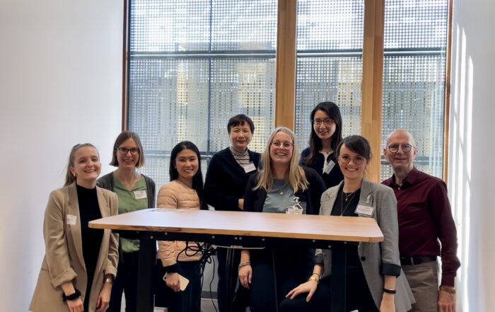Eine Gruppe von acht Personen der Hochschule Coburg lächelt und posiert drinnen vor einem großen Fenster. Sie stehen um einen Stehtisch mit transparenter Plattform herum, vor einem Holzboden und hellem Licht, und verkörpern einen Moment der Kameradschaft und Innovation.