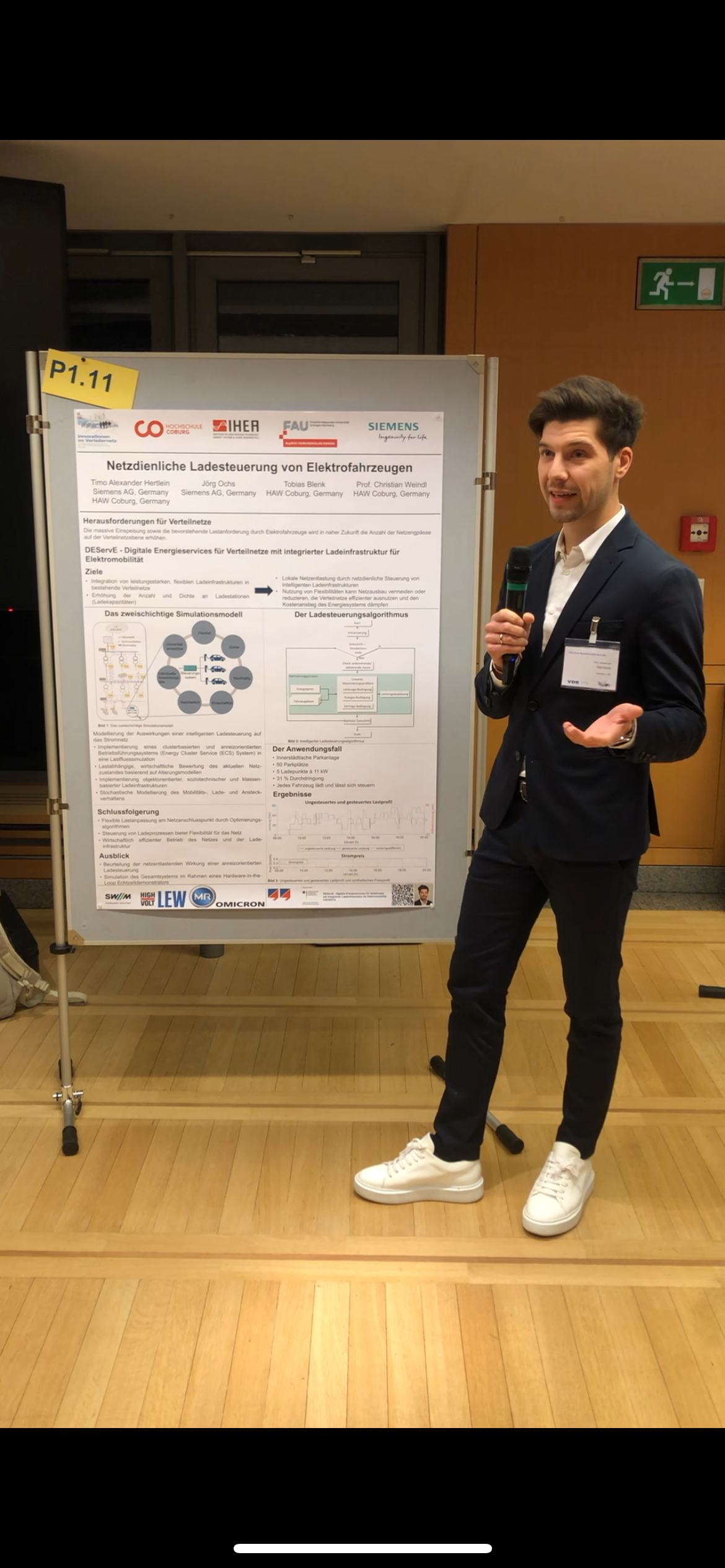 Eine Person im Anzug präsentiert auf einer Konferenz der Hochschule Coburg ein Poster zum Thema Netzmanagement für Elektrofahrzeuge. Sie steht drinnen, hält ein Mikrofon in der Hand und zeigt auf das Poster, das Text, Diagramme und Logos mehrerer Organisationen enthält.