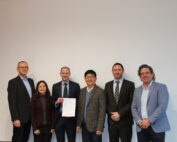 Eine Gruppe von sieben Personen, fünf Männer und zwei Frauen, steht Seite an Seite in Business-Kleidung an der Hochschule Coburg. Ein Mann hält ein Dokument in der Hand. Sie posieren vor einer schlichten weißen Wand.