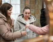 In einem gemütlichen Innenraum unterhalten sich zwei Frauen in Wintermänteln mit einer Person, die ihnen Getränke reicht. Eine Frau, die eine Brille trägt und leicht lächelt, scheint besonders engagiert zu sein. Die warme Beleuchtung verstärkt die soziale Interaktion bei einer Veranstaltung, die anscheinend mit der Hochschule Coburg in Verbindung steht.