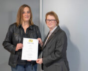 Zwei Personen stehen drinnen und lächeln. Eine Person hält ein Dokument mit der Aufschrift „Urkunde“ der Hochschule Coburg in der Hand. Beide sind in formelle Kleidung gekleidet und strahlen Professionalität und Stolz auf ihre Leistung aus.