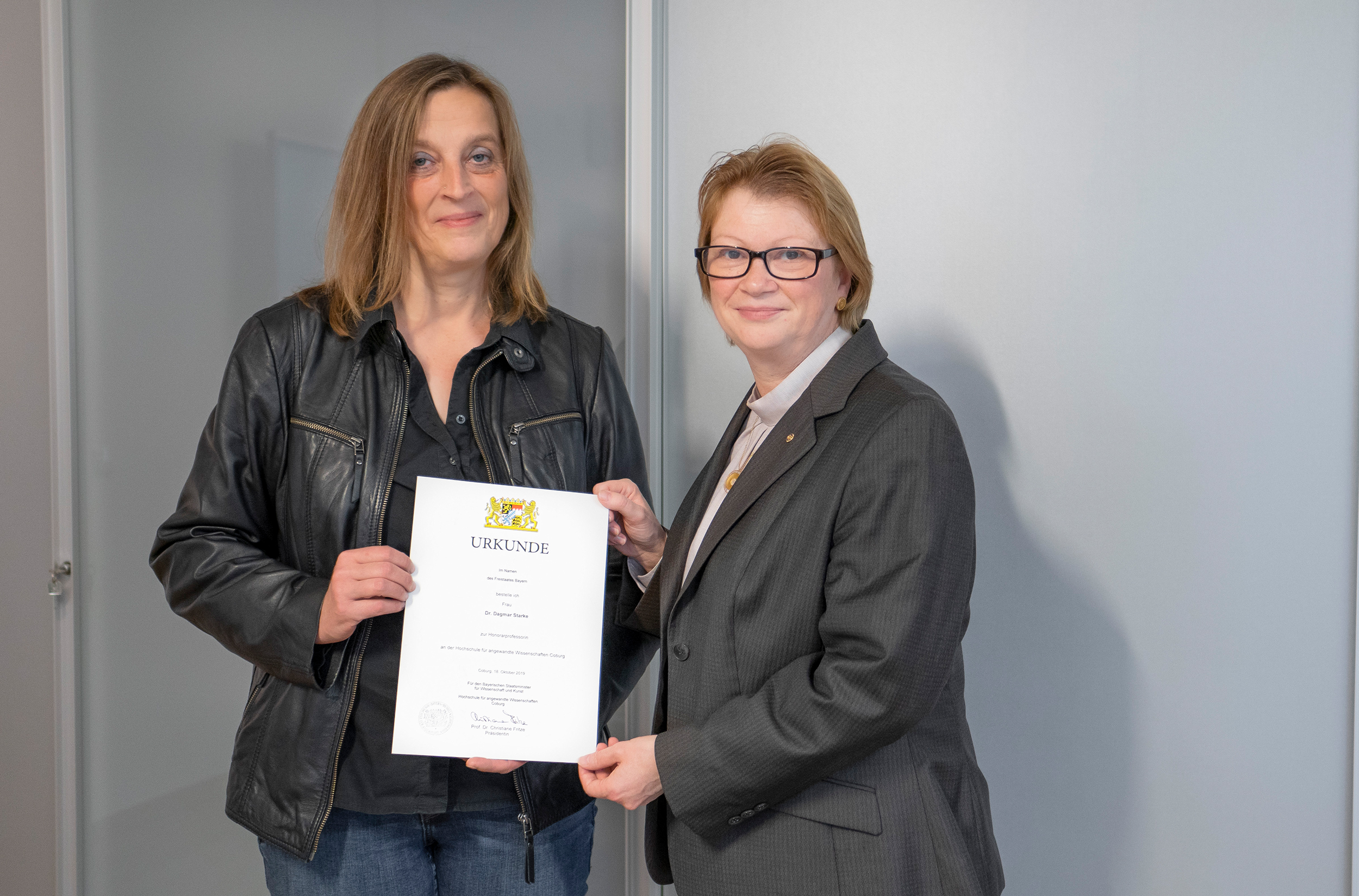 Zwei Personen stehen drinnen und lächeln. Eine Person hält ein Dokument mit der Aufschrift „Urkunde“ der Hochschule Coburg in der Hand. Beide sind in formelle Kleidung gekleidet und strahlen Professionalität und Stolz auf ihre Leistung aus.