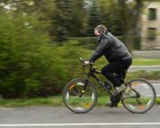 Eine Person mit schwarzer Jacke und Mund-Nasen-Bedeckung fährt auf einem schwarzen Fahrrad zügig an der Hochschule Coburg vorbei. Die grünen Büsche und der unscharfe Hintergrund betonen ihre schnelle Bewegung.