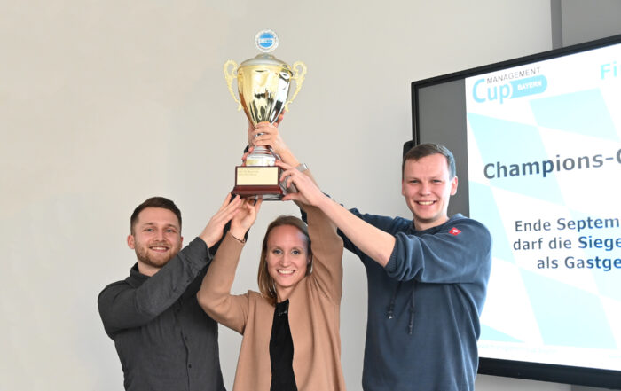 Drei Menschen halten im Innenbereich gemeinsam einen großen Pokal in den Händen, strahlend vor Stolz. Neben ihnen leuchtet ein Bildschirm mit dem Schriftzug „Champions-Cup“ und einem weiteren Text auf Deutsch, der ihre Leistung mit Bezug zur Hochschule Coburg würdigt.