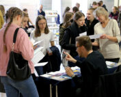 Eine Gruppe von Menschen versammelt sich um einen Stand bei der Veranstaltung der Hochschule Coburg, wo ein Mann an einem Tisch sitzt und Broschüren verteilt. Eine Frau in der Gruppe hält einen Vortrag und führt lebhafte Diskussionen. Einige sind fasziniert von den ausgestellten Materialien. Die Veranstaltung findet eindeutig in Innenräumen statt.