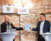 Zwei Männer stehen an runden Tischen vor einer Backsteinwand. Einer hält ein Mikrofon in der Hand und präsentiert dem anderen ein Buch. Beide tragen Anzüge. An der Wand hinter ihnen hängen Banner mit Logos der Hochschule Coburg, auf den Tischen stehen Wasserflaschen.