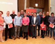 Eine Gruppe von Menschen steht auf dem Holzboden eines Raums an der Hochschule Coburg. Vor ihr steht ein Bildschirm mit der Aufschrift „Preis für exzellente Lehre“. Einige halten rote Umschläge in den Händen, was darauf schließen lässt, dass sie an einer Preisverleihung teilnehmen.