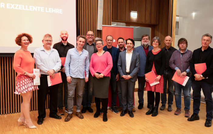 Eine Gruppe von Menschen steht auf dem Holzboden eines Raums an der Hochschule Coburg. Vor ihr steht ein Bildschirm mit der Aufschrift „Preis für exzellente Lehre“. Einige halten rote Umschläge in den Händen, was darauf schließen lässt, dass sie an einer Preisverleihung teilnehmen.